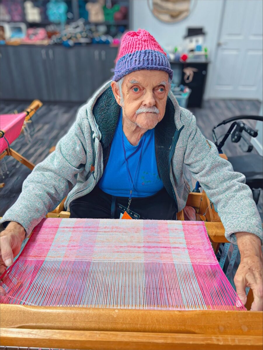 An adult with developmental disabilities creates a vibrant fabric piece on a loom in the weaving program at Rainbow Acres in Camp Verde, Arizona. The workspace is filled with colorful threads, showcasing the creativity and focus of the individual.