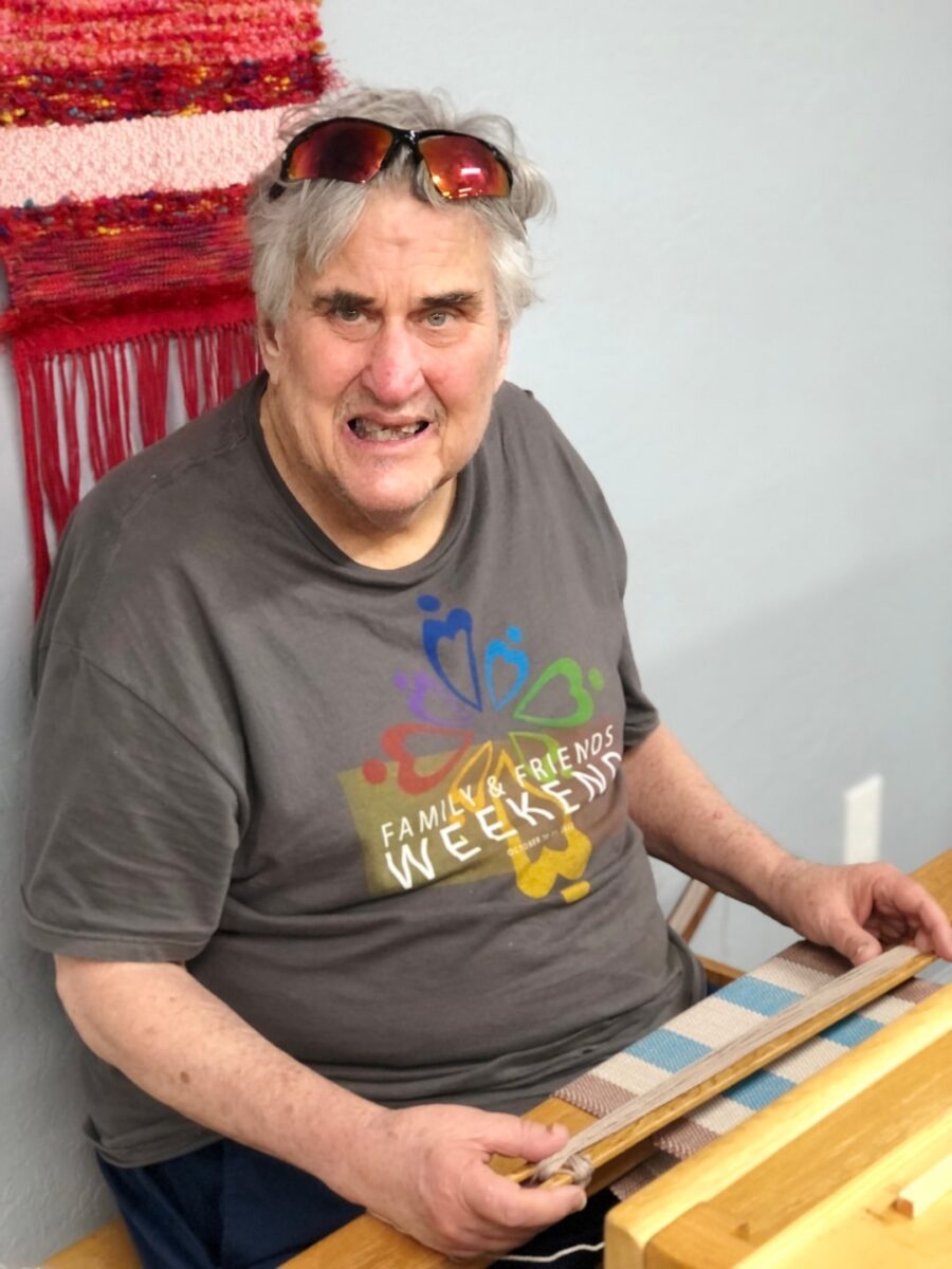 An adult with developmental disabilities focuses on weaving a piece of fabric in the creative arts weaving program at Rainbow Acres in Camp Verde, Arizona. The individual is seated at a loom, their hands carefully guiding threads through the intricate design. Bright natural light fills the room, highlighting the colorful yarns and the supportive, engaging environment.