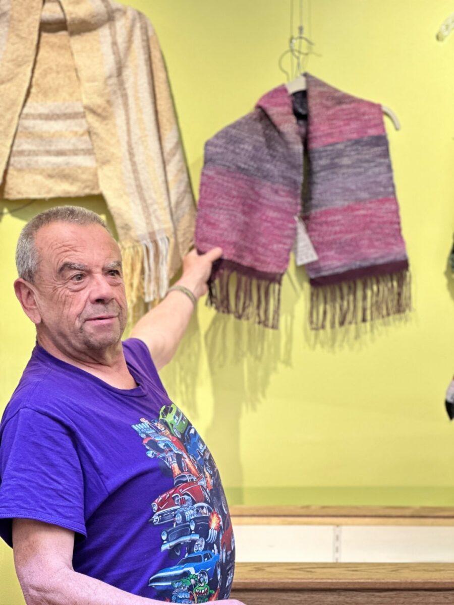 An individual at Rainbow Acres participates in the creative arts program, weaving a piece of fabric on a loom. The image highlights their concentration and the vibrant threads being used in the design.